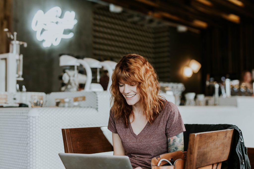 A smiling woman sits in a coffee shop with laptop in lap. This could represent online therapy in California. An online therapist can help with online depression therapy and other services.